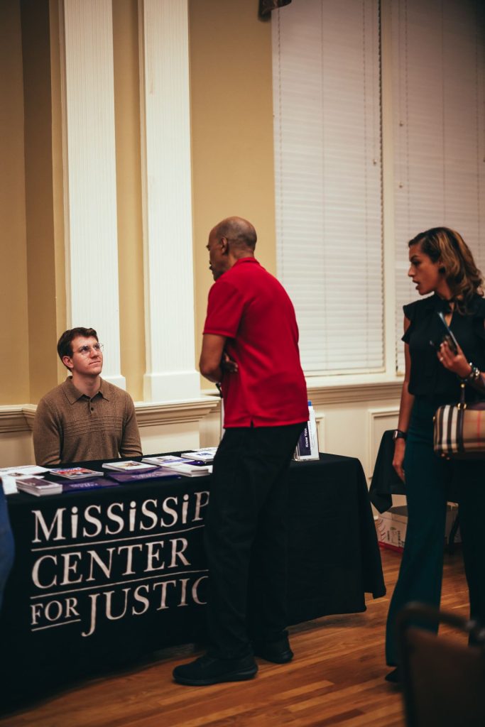 Gavin listens to a man share his experience while tabling at a Homeowner Empowerment Seminar in Hattiesburg, MS. A woman passing by looks inquisitively at the table replete with informational brochures.